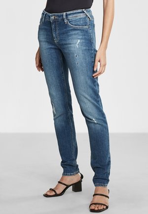 Jeans de mezclilla azul de corte slim con detalles desgastados, diseño de cinco bolsillos y cierre de botón. Llevados con sandalias de tacón negras.