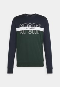 Mörkgrön och marinblå långärmad sweatshirt med tydlig "SPORT" text och ett vitt horisontellt band. Mjuk tygkvalitet och åtsittande mudd.