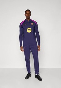 Vyras stovi priešais paprastą foną, dėvėdamas tamsiai mėlyną ir violetinę FC Barcelona treniruočių kostiumą su geltonais logotipais ir juodus sportbačius.