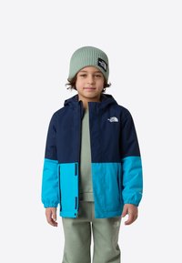 Jachetă albastră și bleumarin cu glugă, prevăzută cu închidere cu fermoar și manșete elastice. Poartă un hanorac verde deschis și o căciulă verde cu model reiat.