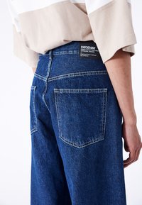 Dunkelblaue Denim-Jeans mit weitem Bein, sichtbaren Nähten, zwei Gesäßtaschen und einem Markenlabel am Bund, das über eine weiche Textur verfügt.