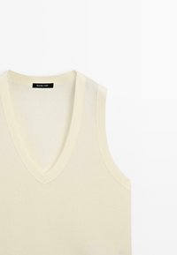 Ljusbeige ärmlös topp med V-ringning, tillverkad av ett slätt tyg, med en subtil textur och en svart logotag inuti ringningen.
