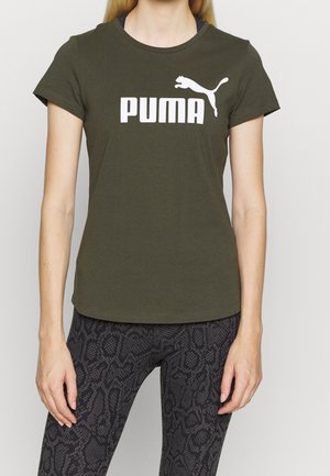 Persoon draagt een olijfgroene Puma t-shirt en zwarte legging met patroon, staand tegen een effen achtergrond.