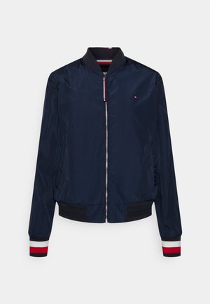 Marineblauwe bomberjack met rits aan de voorkant, geribde kraag en manchetten met rood-wit-zwarte strepen, klein logo op de borst.