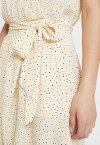 Robe jaune avec un motif à pois blancs et noirs, présentant une taille froncée et une ceinture nouée. Le tissu a un aspect doux et texturé.