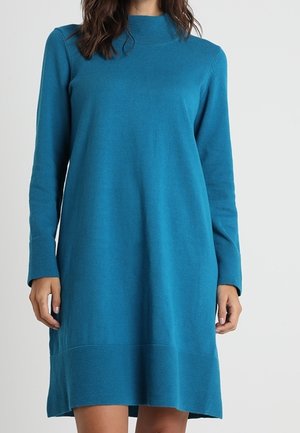 Femme portant une robe en maille bleu sarcelle à manches longues, longueur genou, avec des poignets et un ourlet côtelés, debout devant un fond clair uni.