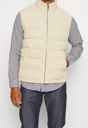 Mand iført beige quiltet ærmeløs vest over sort-hvid ternet skjorte og mørke jeans, med den ene hånd i vestens lomme, stående foran en hvid baggrund.
