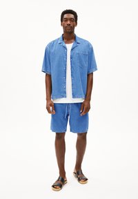 Denim overhemd met korte mouwen, lichtblauwe kleur, voorzak; bijpassende blauwe denim shorts, platte zwarte sandalen. Eenvoudige, casual outfit.