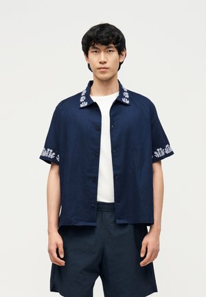 Jeune homme portant une chemise à manches courtes bleu marine avec des broderies florales blanches sur le col et les manches, associée à un short bleu marine et un t-shirt blanc.