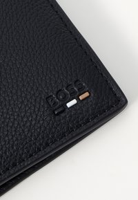 Portefeuille en cuir noir avec une surface texturée, présentant le logo "BOSS" en relief et des accents colorés en noir, blanc et marron.