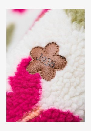 Fuzzy deka s teksturiranom bijelom površinom koja ima ružičaste i zelene uzorke. Na njoj je pričvršćen smeđi kožni tag u obliku cvijeta s natpisom "JoJo".