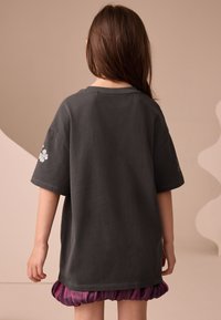 Grå bomullst-shirt med korta ärmar, med blommig broderi på vänster ärm. Fållen är längre baktill, bärs med en lila kjol.