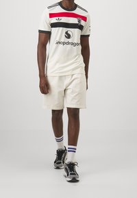 Valkoinen urheilupaita, jossa on punaisia ja mustia vaakaraitoja, mustareunus ja logo. Yhdistetty beigen värisiin shortseihin ja mustiin tennareihin, joissa on valkoisia yksityiskohtia.