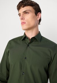 Cămașă butonată verde închis, dintr-un material neted, având un guler clasic și nasturi negri, cu mâneci lungi adaptate pentru un aspect ajustat.