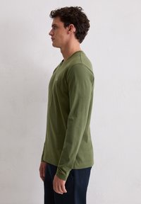 Koszulka z długim rękawem w kolorze oliwkowej zieleni, wykonana z bawełny, z okrągłym dekoltem i dopasowanym krojem. Minimalistyczne logo na piersi.