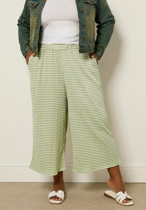 Persona con pantalones anchos de cuadros en verde claro y blanco, sandalias blancas, camiseta blanca y chaqueta vaquera verde descolorida, de pie contra una pared beige.