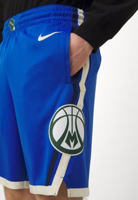 Pantalones cortos deportivos azules de tejido de malla con rayas negras y crema, que presentan un gran logotipo de baloncesto verde y blanco en la pierna izquierda.