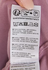 Plejeetiket på en lys lilla baggrund, der viser tekst på flere sprog. Nøgleoplysninger: 95% bomuld, 5% elastan.