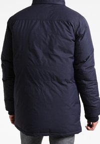 Marinblå vadderad jacka med hög krage, med en slät yta, elastiska ärmslut och en längre passform, utformad för värme och komfort.