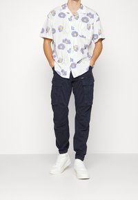 Kortärmad skjorta med vit bakgrund och lila blommönster, parat med marinblå cargobyxor och vita sneakers.