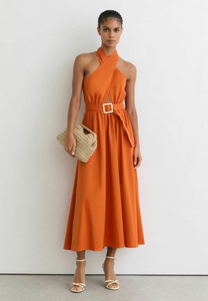 Femme portant une robe orange sans manches à col montant avec une ceinture, tenant une pochette beige tissée, debout contre un mur blanc.
