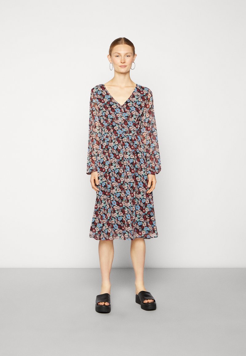 Vestido floral com decote em V profundo, mangas longas e bainha evasê. Apresenta um fundo escuro com flores coloridas. Combinado com sandálias pretas de plataforma.