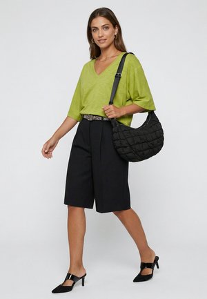 Nainen, joka käyttää limevihreää V-kaula-aukkoinen toppia, mustia polvipituisia shortseja, mustia korkokenkiä ja kantaa mustaa tikattua olkalaukkua.