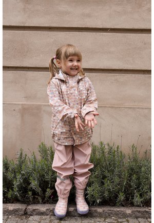 Nepremokavá ružová dažďová bunda s motívmi srdiečok, k nej ladia ružové nepremokavé nohavice a gumáky so vzorom. Dieťa stojí pri zelenej rastlinnej výzdobe.