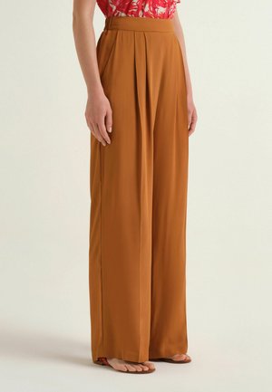 Pantalon large couleur rouille à plis à l'avant, porté par une personne avec un top rouge à motifs et des sandales plates, debout devant un fond uni.