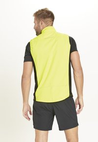 Leichte, neon-gelbe Weste aus atmungsaktivem Material, mit hohem Kragen, schwarzen Seitenpaneelen und einem dezenten Herzschlaglinien-Design.