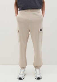 Beige Sweatpants mit elastischem Bund und umgeschlagenen Säumen, ausgestattet mit zwei seitlichen Taschen und einem Patch-Label am linken Oberschenkel.