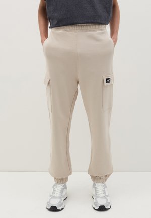 Pantalones de chándal beige con cintura elástica y dobladillos ajustados, que presentan dos bolsillos laterales y una etiqueta de parche en el muslo izquierdo.