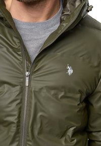 Olivinės spalvos pūkinė striukė su blizgiu paviršiumi, užtrauktuku ir mažuoju baltu logotipo ženklu. Dėvima ant pilkos megztinės su iškilimais apykakle.