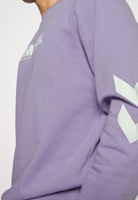Světle fialová mikina vyrobená z bavlny. Na přední straně se nachází bílé logo a na rukávu bílé geometrické zdobení. Měkká textura.