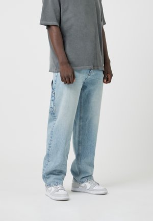 Personne portant un jean large bleu clair, un T-shirt gris surdimensionné et des baskets blanc-gris, debout devant un fond clair uni.