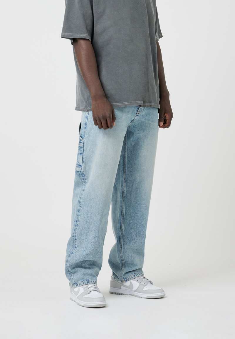 Personne portant un jean large bleu clair, un T-shirt gris surdimensionné et des baskets blanc-gris, debout devant un fond clair uni.