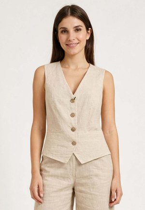 Donna con lunghi capelli castani che indossa un gilet beige senza maniche con bottoni e pantaloni beige abbinati, sorridente e rivolta verso di fronte.