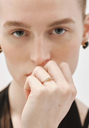 Mujer de piel clara y ojos azules sostiene la mano cerca de la boca, lleva un delgado anillo de oro con pequeños diamantes en el dedo.