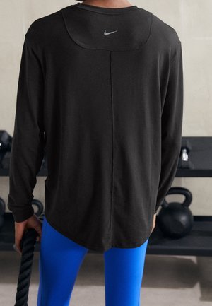 Person iført sort langærmet Nike-shirt og blå leggings, der holder et battle rope i et fitnesscenter med kettlebells på hylderne i baggrunden.