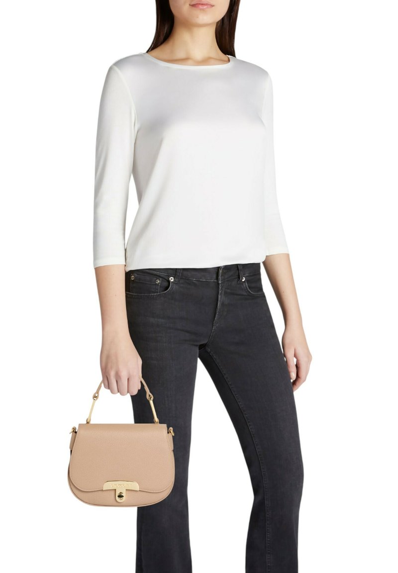 Borsa in pelle beige con una forma curva, dettagli in oro e manico superiore, tenuta da una persona con indosso un top bianco a maniche lunghe e jeans neri.