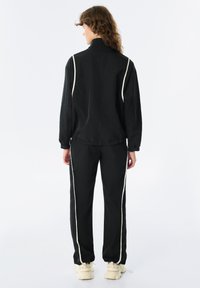 Tuta sportiva nera con colletto alto, dotata di strisce laterali bianche; i pantaloni sono a vestibilità ampia con polsini svasati, realizzati in tessuto leggero.