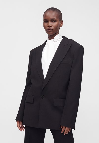 Femme aux cheveux courts portant un blazer noir oversize et un chemisier blanc à col haut sur un fond uni.