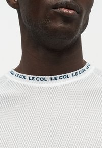 Gros plan du cou d'un homme portant une chemise en mesh blanche avec un col répétant le texte "LE COL" en bleu foncé.