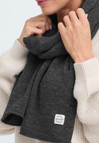 Mørkegrå strikket skjerf med en myk tekstur. Har et logo-patch merket "OXMO" i den ene enden. Båret rundt halsen over en lys genser.