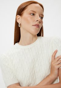 Suéter de punto blanco con mangas cortas, cuello redondo y sutiles detalles de textura. La modelo lleva pendientes de aro plateados, con el cabello largo y liso.