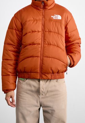 Veste matelassée orange de The North Face fermée, portée avec un pantalon beige, main gauche dans la poche de la veste et main droite détendue sur le côté.