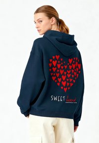 Sweat à capuche surdimensionné bleu marine avec un design de cœur rouge composé de cœurs plus petits au dos, avec le texte "SWEET heart" en blanc.
