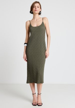 Donna che indossa un vestito midi verde oliva testurizzato con spalline sottili, tacchi neri con cinturino alla caviglia, orecchini neri e due braccialetti al polso sinistro.