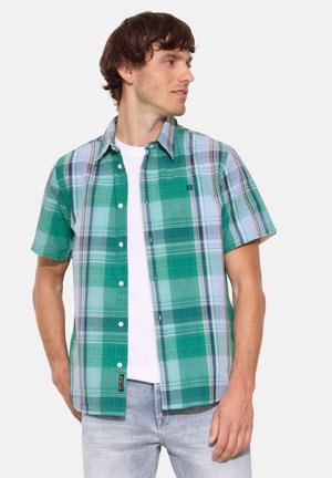 Mann trägt grün-blau kariertes kurzärmeliges Hemd über weißem T-Shirt mit hellen Jeans, schaut nach rechts.