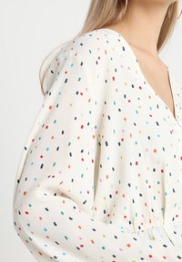 Blusa leggera in crema con scollatura a V profonda, maniche lunghe e motivo a pois multicolore. Vestibilità ampia con dettaglio in vita elastico.